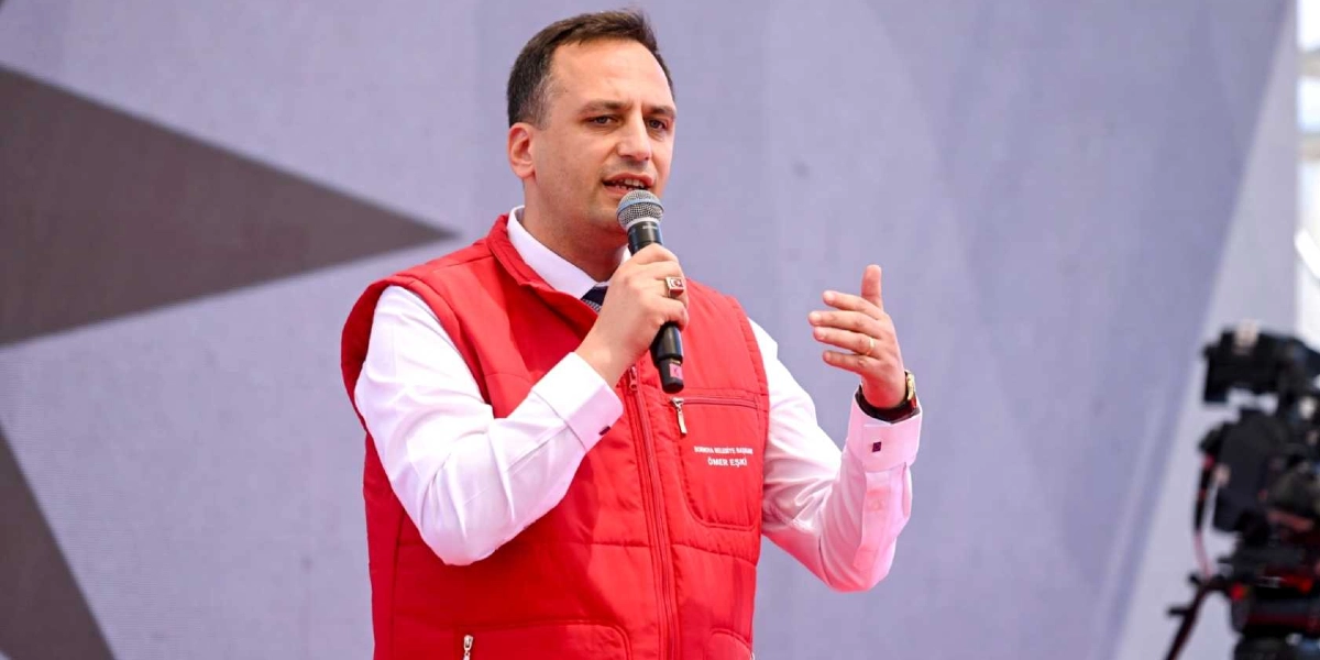 Bornova Belediye Başkanı Ömer Eşki, beyaz gömlek ve üzerine giydiği kırmızı bir yelekle, elinde mikrofon tutarak konuşma yapıyor. Yeleğinin üzerinde 