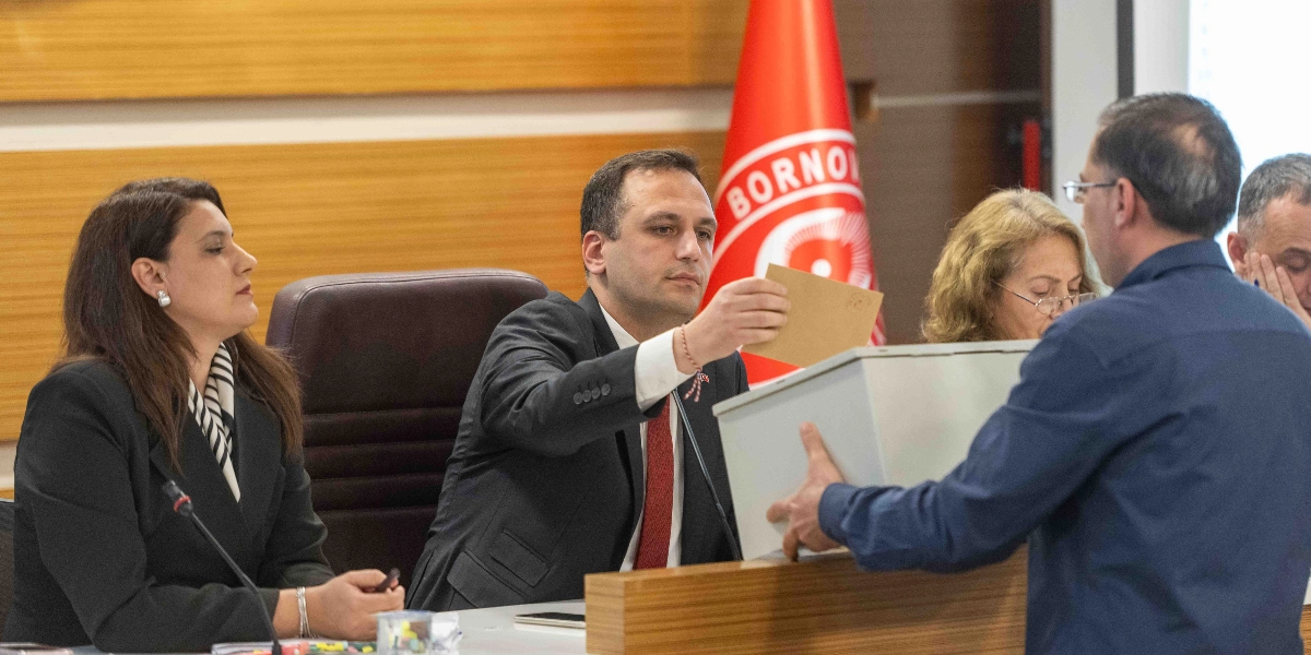 Bornova Belediyesi meclis salonunda, takım elbiseli bir erkek üye elindeki oy zarfını sandığa atıyor. Masanın diğer tarafında, yanında siyah ceketli bir kadın üyenin oturduğu bu sırada, arka planda Bornova Belediyesi'nin kırmızı renkli logosunun bulunduğu bayrak yer alıyor.
