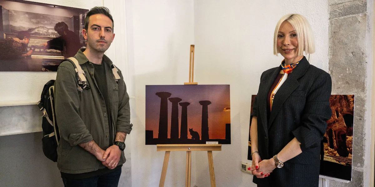 Bir müze veya sergi alanında, ortadaki ahşap şövale üzerinde duran ve gün batımında antik sütunlar ile bir kedinin silüetini gösteren fotoğrafın iki yanında duran bir erkek ve bir kadın. Sol taraftaki erkek, haki renkli bir gömlek ve sırt çantasıyla dururken, sağ taraftaki sarı saçlı kadın siyah bir ceket ve boynunda renkli bir fularla poz veriyor. Arka plandaki duvarlarda başka sergi fotoğrafları da yer alıyor.