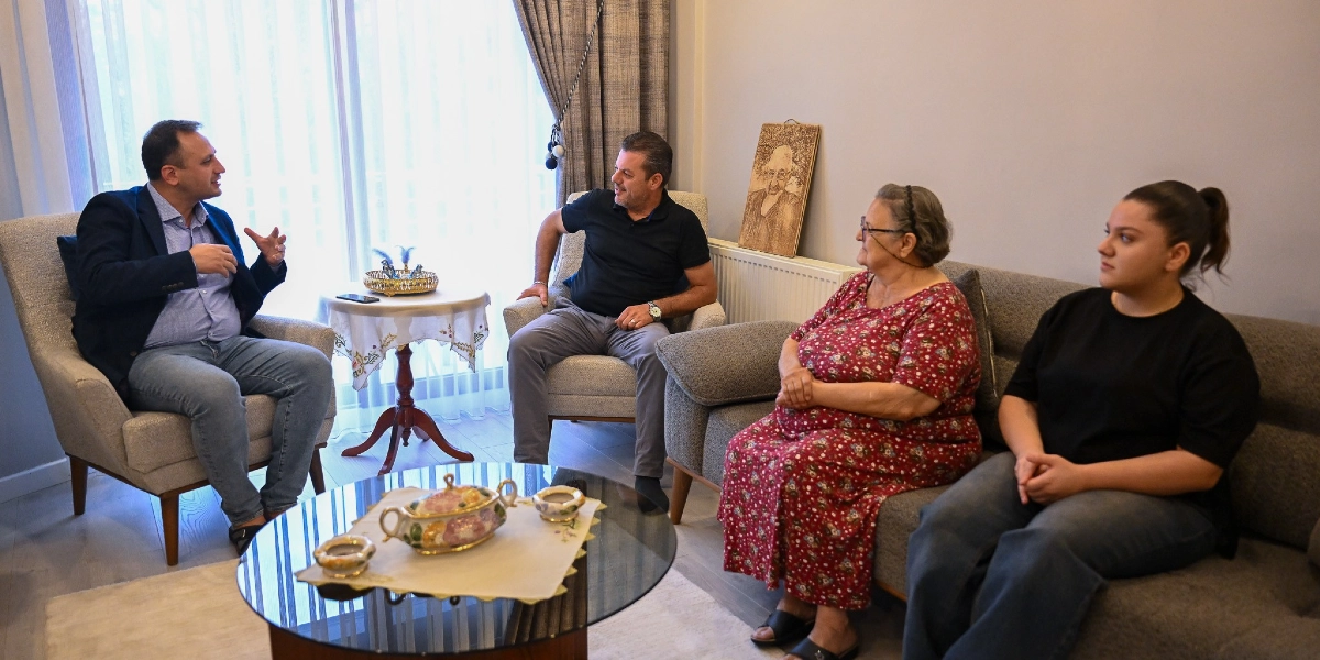 Bornova Belediye Başkanı Ömer Eşki, bir ev ziyaretinde yaşlı bir kadın ve ailesiyle oturma odasında sohbet ediyor. Başkan Eşki, mavi gömlek ve lacivert ceket giymiş şekilde tekli koltukta otururken, karşısında oturan aile üyeleriyle konuşuyor. Odanın köşesinde duvara yaslanmış ahşap üzerine işlenmiş bir portre tablosu bulunuyor.
