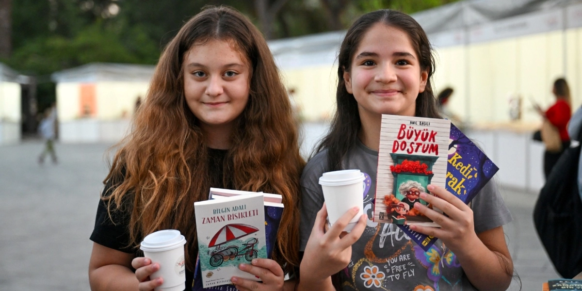 Açık havada düzenlenen bir kitap fuarı alanında, ellerinde kitaplar ve karton kahve bardakları tutan, gülümseyen iki genç kız. Soldaki kız elinde Bilgin Adalı'nın Zaman Bisikleti kitabını, sağdaki kız ise Anıl Basılı'nın Büyük Dostum kitabını tutuyor. Arka planda fuar stantları ve bulanık bir insan kalabalığı görülüyor.
