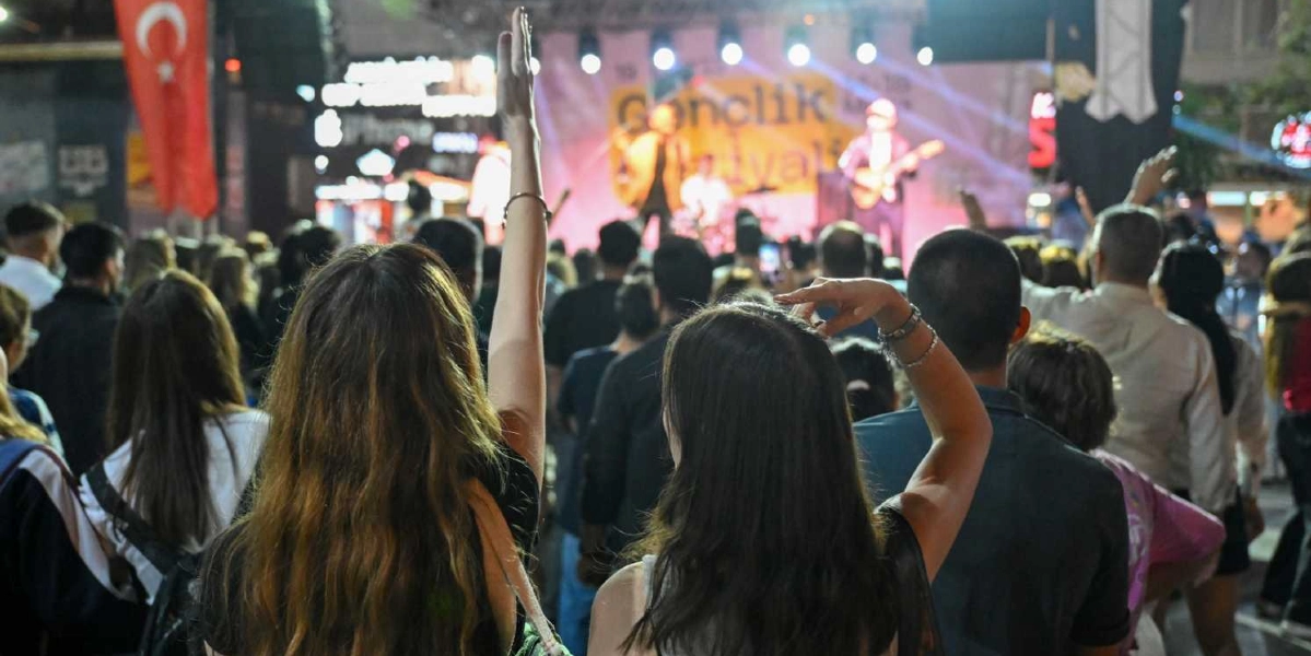 Açık havada düzenlenen bir gençlik festivalinde sahnedeki müzisyenleri izleyen kalabalık bir grup insan. Ön planda, ellerini havaya kaldırmış konser coşkusunu yaşayan gençler görülüyor. Sahne üzerinde ışıklandırmalar ve üzerinde 