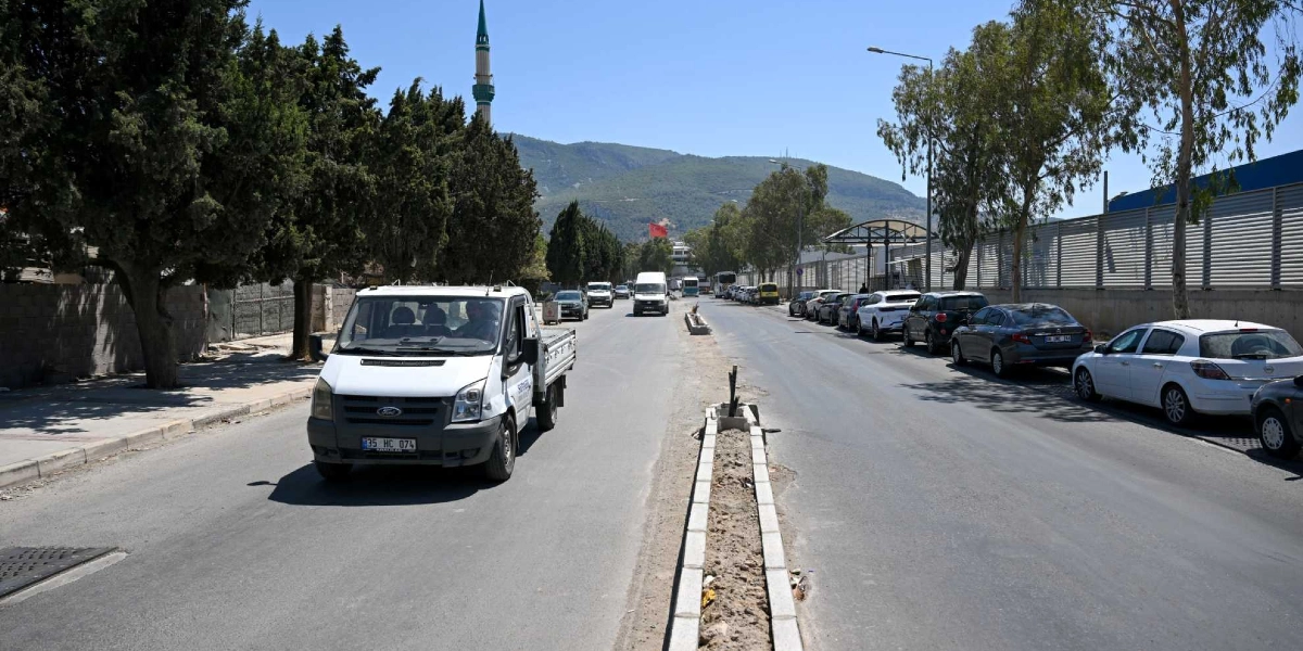 Bornova'da ağaçlıklı bir yolun ortasında, üzerinde çalışma yapılan bir refüj ve kenarlara park etmiş araçların bulunduğu caddede ilerleyen beyaz bir kamyonet görülüyor. Arka planda dağ manzarası ve bir cami minaresi yer alıyor.