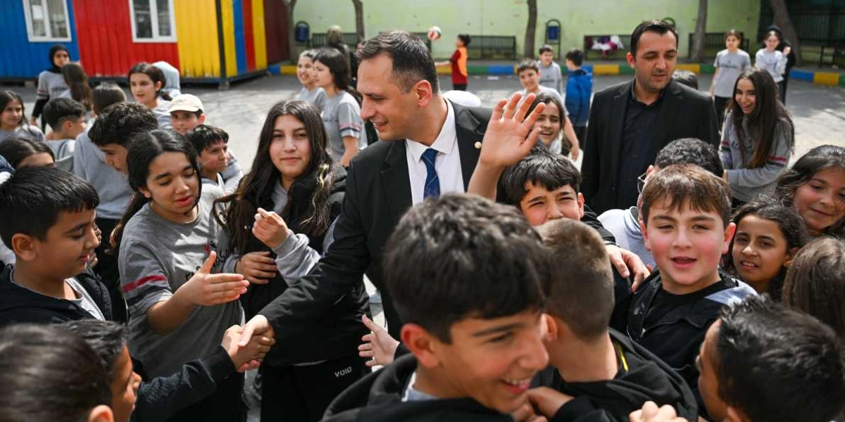 Takım elbiseli ve kravatlı bir adam, konteyner kent benzeri bir alanda bir grup çocuk ve gençle bir araya gelerek onlarla tokalaşıyor ve el sallıyor. Çevresini saran çocuklar gülümseyerek kendisiyle etkileşim kuruyor, arka planda ise diğer çocuklar oyun oynuyor.
