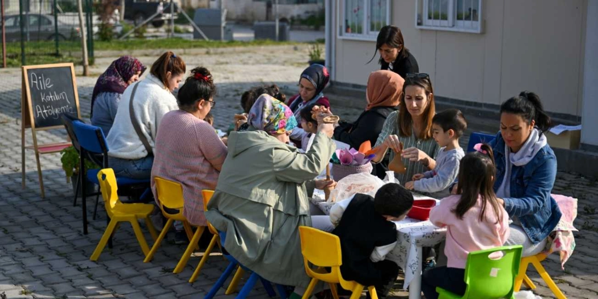 Açık havada, beyaz bir masa etrafında toplanmış anneler ve çocukları çim adam yapımı etkinliğine katılıyor. Masanın başında üzerinde 