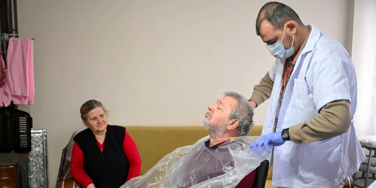 Beyaz önlüklü, maskeli ve eldivenli bir sağlık görevlisi, ev ortamında oturan yaşlı bir adamın üzerine koruyucu plastik bir örtü yerleştiriyor. Yan taraftaki koltukta oturan orta yaşlı bir kadın ise bu süreci izliyor.