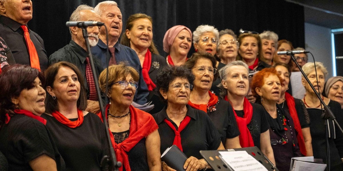 Siyah kıyafetler giymiş, boyunlarında kırmızı fularlar takan ve ellerinde notalar tutan ileri yaştaki bir koro grubu, sahnede mikrofonlar eşliğinde şarkı söylüyor.