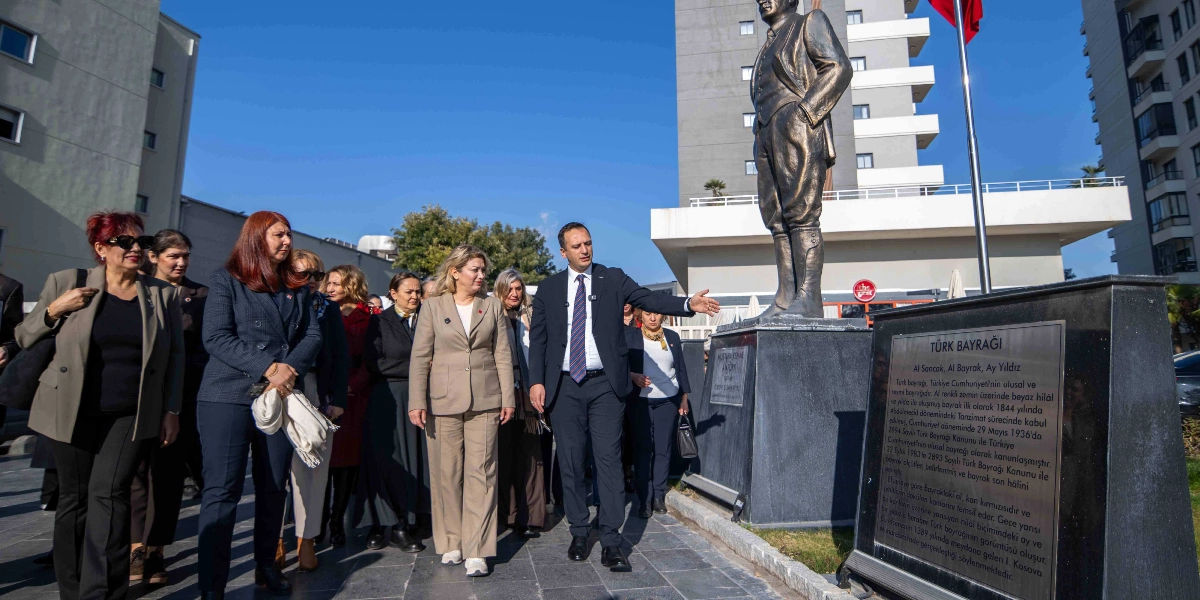 CHP Kadın Kolları Genel Başkanı Asu Kaya ve beraberindeki heyet, Bornova Belediye Başkanı Ömer Eşki ile birlikte Atatürk heykeli önünde duruyor. Başkan Eşki, elini heykelin yanındaki Türk bayrağı hakkında bilgi veren plakete doğru uzatarak heyete açıklamalarda bulunuyor. Açık havada, güneşli bir günde gerçekleşen ziyarette grup, heykelin bulunduğu meydanda bir arada yer alıyor.