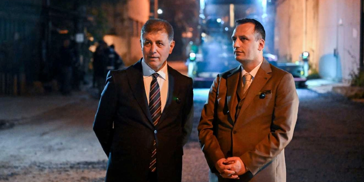 Koyu renk takım elbiseli ve çizgili kravatlı bir erkek ile yanında açık kahverengi takım elbiseli ve açık renk kravatlı başka bir erkek, gece vakti dış mekanda yan yana durarak ileriye doğru bakıyor. Arka planda ise hafif bulanık bir şekilde çalışma alanını aydınlatan ışıklar ve iş makineleri seçiliyor.