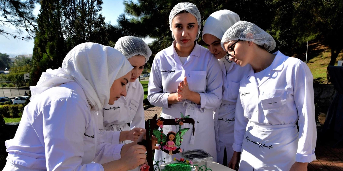 Beyaz önlük ve bone giymiş beş genç kadın, dış mekanda bir masa etrafında toplanmış, üzerinde peri figürlü bir salıncak bulunan pastayı inceliyor. Kadınların önlüklerinin üzerinde 