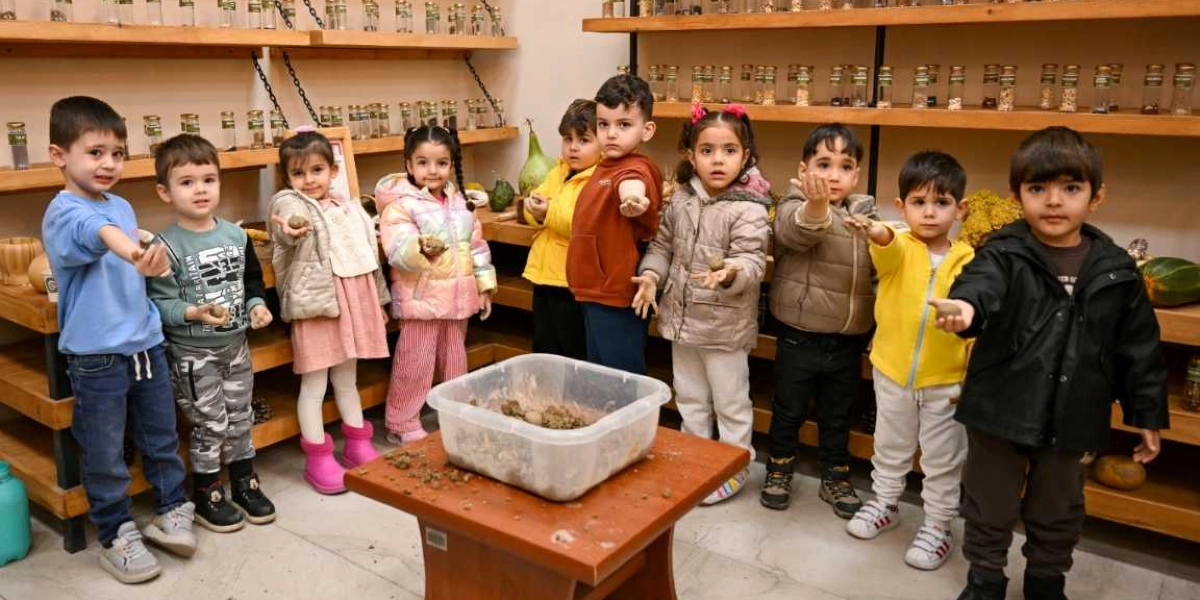 Ahşap raflarla çevrili bir odada, önlerindeki masada bulunan plastik bir kaptan aldıkları toprak parçalarını avuçlarında tutarak kameraya gösteren dokuz küçük çocuk yan yana duruyor. Çocuklar kışlık kıyafetler giymiş durumda ve arka plandaki raflarda cam kavanozlar içinde çeşitli tohumlar sergileniyor.