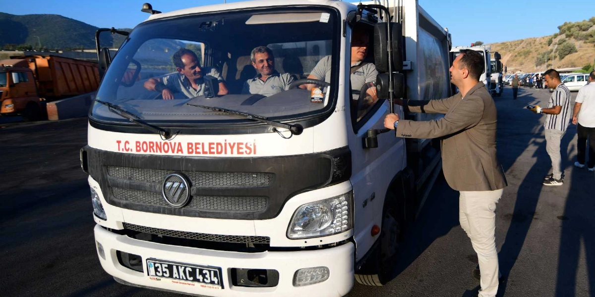 Üzerinde T.C. Bornova Belediyesi yazan beyaz bir çöp kamyonunun şoför mahallinde üç belediye çalışanı oturuyor. Kamyonun kapısının yanında duran ceketli bir kişi, şoför koltuğundaki çalışanla sohbet ediyor. Arka planda açık havada park halindeki diğer iş araçları ve ayakta duran belediye personelleri görülüyor.