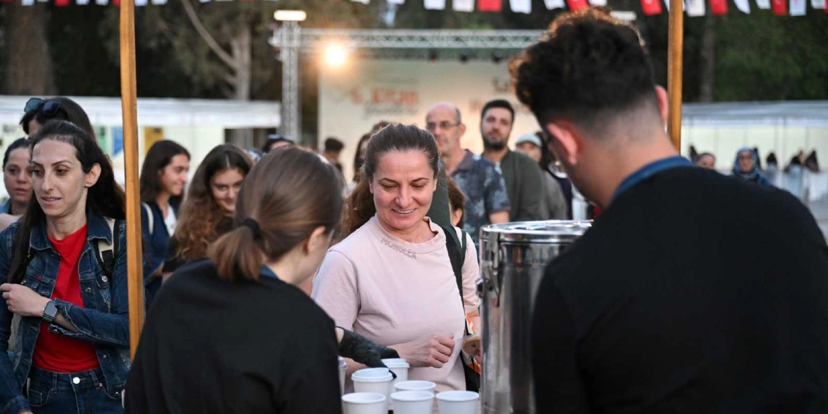 Açık havada düzenlenen bir etkinlikte, siyah giyimli bir görevlinin içecek servisi yaptığı stant önünde bekleyen insanlar. Ön planda, açık renkli tişört giyen güler yüzlü bir kadın ve yanında kot ceketli bir başka kadın yer alıyor. Arka planda ise etkinlik alanındaki kalabalık, ışıklandırılmış bir sahne ve üzerinde Türk bayraklarının asılı olduğu bir düzenek görülüyor.