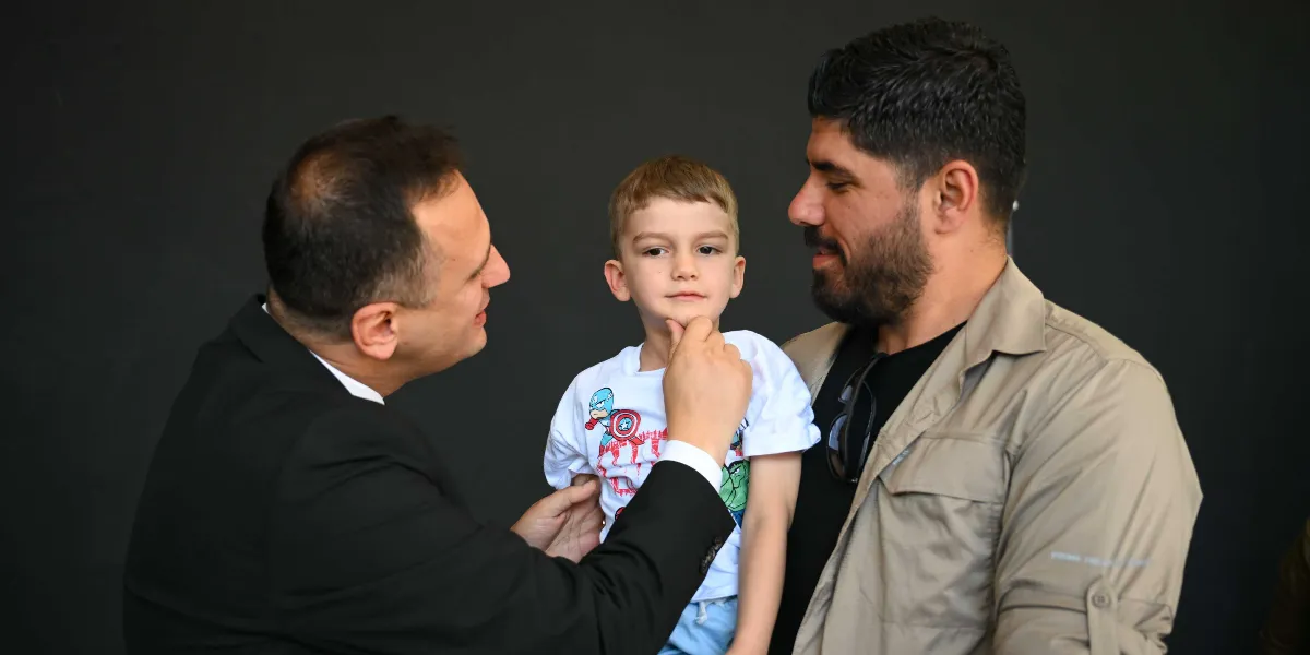 Siyah arka plan önünde, üzerinde süper kahraman baskılı beyaz bir tişört giyen küçük bir erkek çocuğunun çenesini sevgiyle tutan takım elbiseli bir adam ve yanında duran sakallı bir adam.