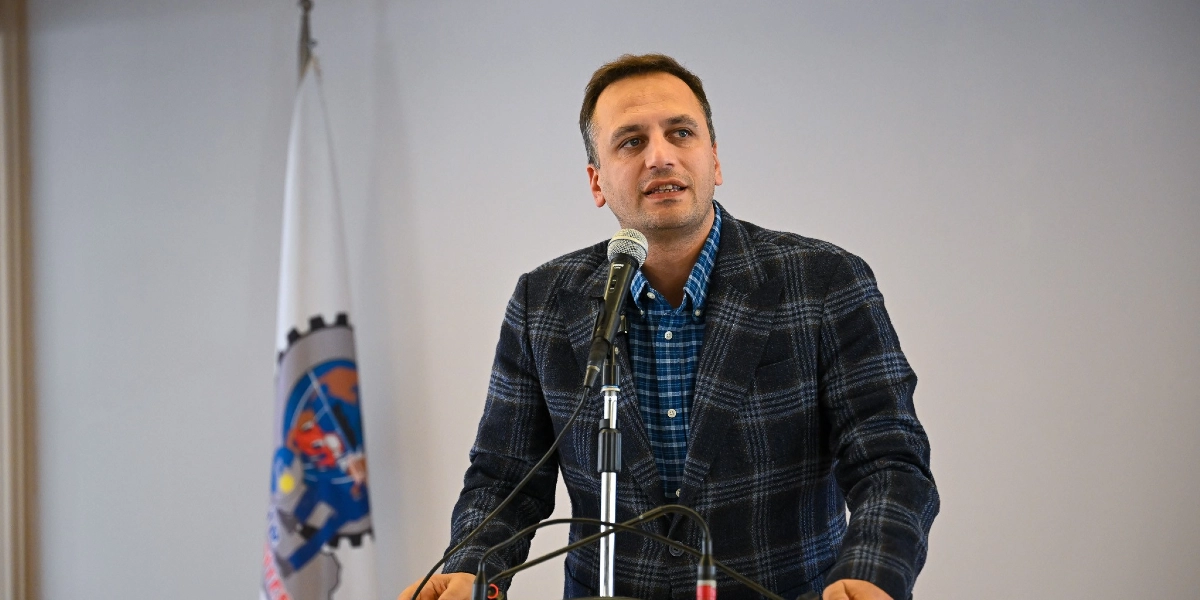 Koyu renkli ekose bir ceket ve mavi kareli bir gömlek giymiş orta yaşlı bir erkek, kürsüde mikrofon başında konuşma yapıyor. Arka planda üzerinde dişli çark figürü bulunan beyaz bir bayrak yer alıyor.