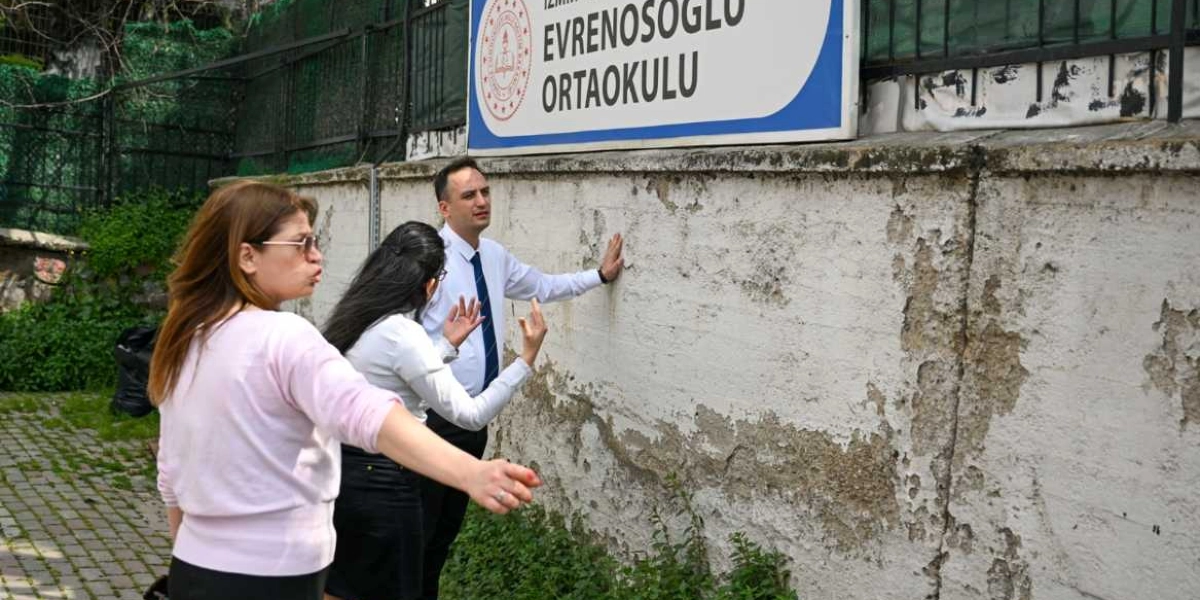 Evrenosoğlu Ortaokulu tabelasının bulunduğu eski ve sıvaları dökülmüş bir duvarın önünde duran üç kişi, elleriyle duvara dokunarak veya işaret ederek hüzünlü bir şekilde çevreyi inceliyor.