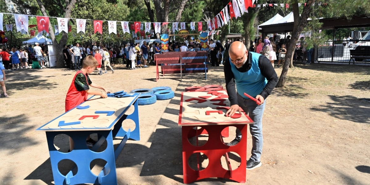 Büyükpark'ta düzenlenen etkinlikte, bir çocuk ve bir yetişkin, üzerlerinde geometrik şekillerin bulunduğu ahşap oyun masalarında vakit geçiriyor. Arka planda ağaçların arasına asılmış Türk bayrakları ve Atatürk posterleri ile süslenmiş alanda, aileler ve çocuklar etkinlik alanında dolaşıyor.