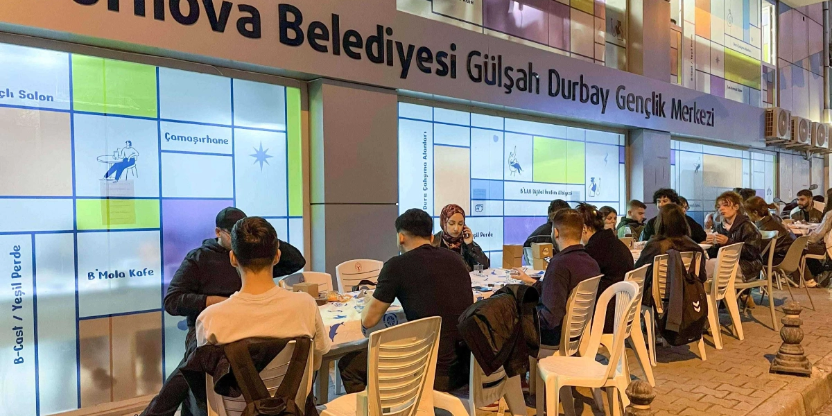 Bornova Belediyesi Gülşah Durbay Gençlik Merkezi binasının önünde, dışarıya kurulan masalarda iftar yapan bir grup genç. Gençler beyaz plastik sandalyelerde oturuyor, masaların üzerinde yiyecek ve içecekler bulunuyor. Binanın dış cephesinde 