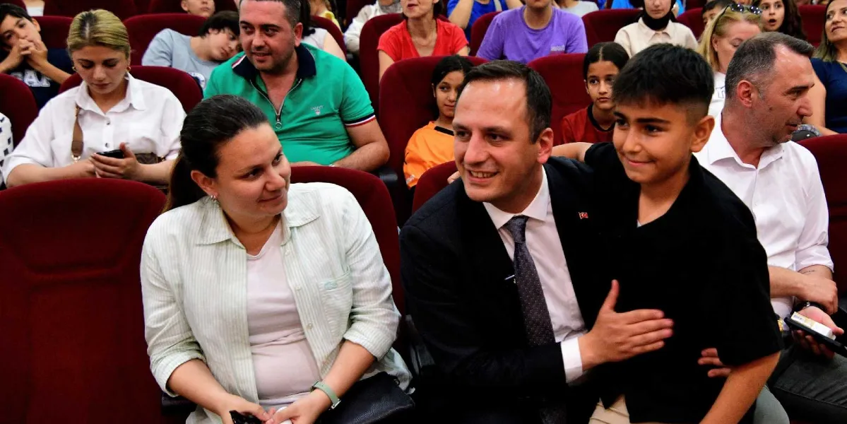 Konferans salonunda kırmızı koltuklarda oturan bir grup insan arasında, takım elbiseli bir erkek, yanındaki genç çocuğun omzuna elini koyarak ona gülümseyerek bakıyor. Çocuğun sol tarafında oturan açık renkli gömlekli bir kadın da onlara doğru bakıyor. Arka planda ise etkinliği izleyen çocuklar ve yetişkinler bulunuyor.