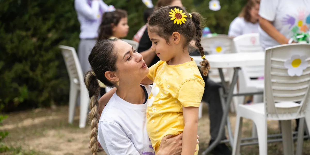 Açık havada, beyaz tişörtlü bir kadın, sarı tişörtlü ve saçında ayçiçeği tokası bulunan küçük bir kız çocuğuna sevgiyle bakarak ona sarılıyor. Arka planda papatya süslemeli beyaz sandalyeler ve etkinlik alanındaki diğer insanlar yer alıyor.
