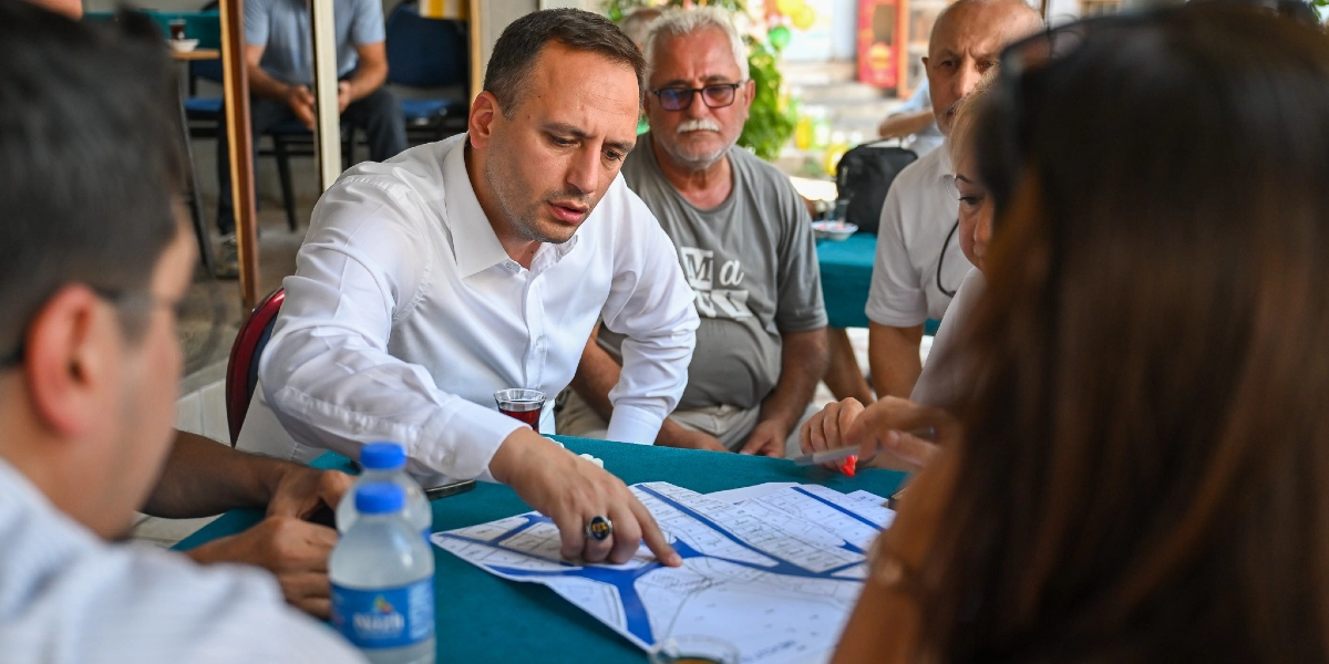 Beyaz gömlekli bir yetkili, bir masanın etrafında toplanan insanlara önündeki teknik bir harita veya proje planı üzerinde işaret ederek bilgi veriyor. Masada su şişeleri ve çay bardakları bulunuyor, çevresinde ise dikkatle dinleyen vatandaşlar yer alıyor.