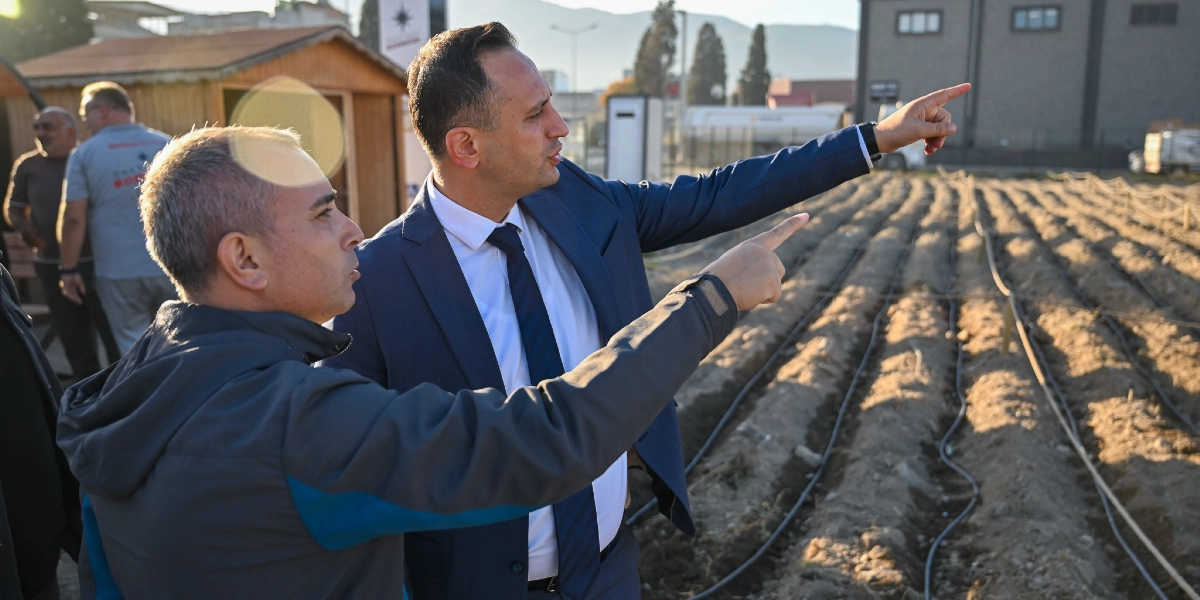 Lacivert takım elbiseli bir yetkili ve yanındaki gri ceketli bir kişi, yan yana durarak elleriyle arazideki tarım alanını işaret ediyor. Arka planda ise damlama sulama sistemi döşenmiş, sürülmüş toprak bir tarla ve uzakta bazı yapılar ile ağaçlar görünüyor.