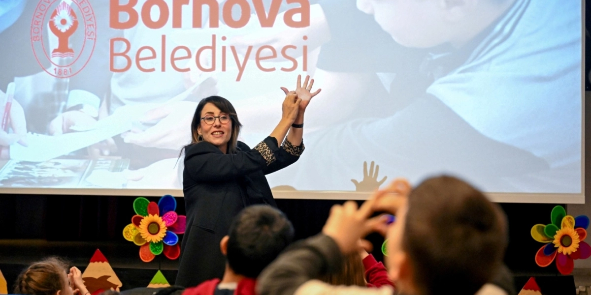 Arka planda Bornova Belediyesi logosunun ve yazısının yer aldığı bir projeksiyon perdesinin önünde, gözlüklü bir kadın eğitmen elleriyle işaret dili kullanarak öğrencilere eğitim veriyor. Ön planda ise eğitmeni izleyen ve elleriyle hareketlere eşlik eden öğrenciler bulunuyor. Sahne dekorunda renkli çiçek figürleri yer alıyor.