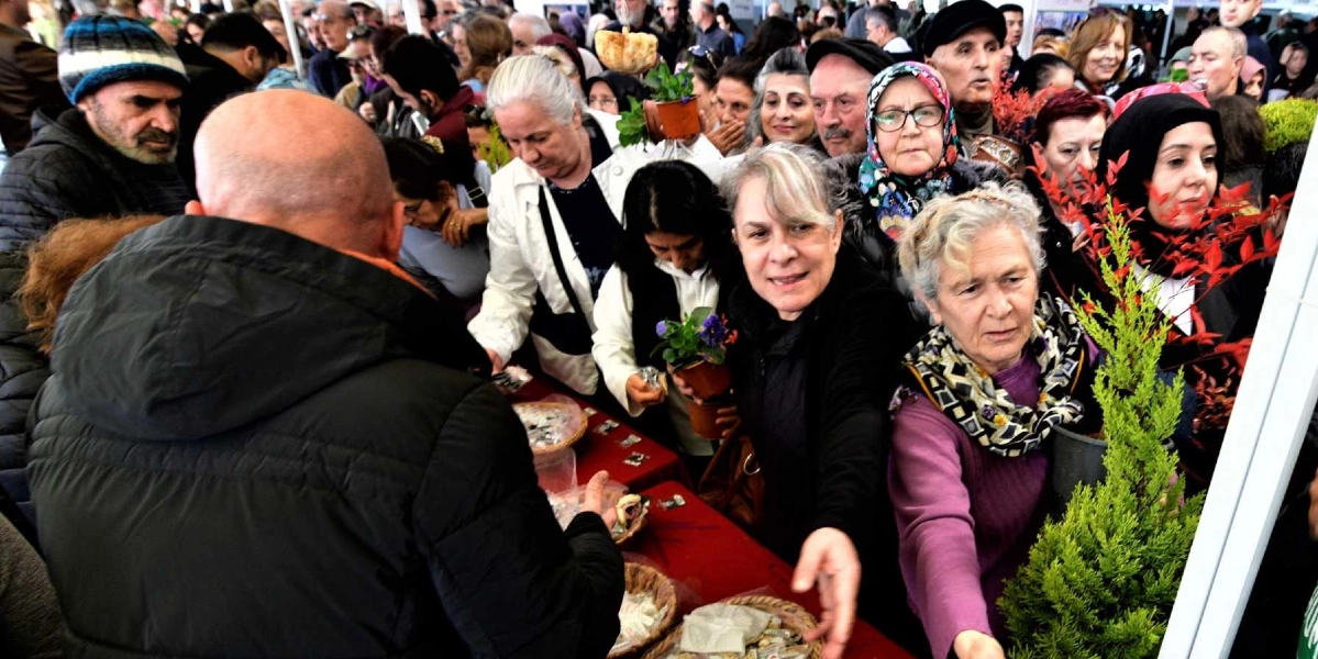 Kalabalık bir grup insanın katıldığı Tohum Takas Şenliği'nde, katılımcılar masaların etrafında toplanmış ellerinde saksı bitkileri ve tohum paketleriyle birbirleriyle etkileşimde bulunuyor. Ön planda, sırtı dönük siyah montlu bir kişi ile karşısındaki kadınlar arasında tohum ve bitki alışverişi gerçekleşiyor. Ortamda farklı yaş gruplarından insanlar, ellerinde çiçekler ve küçük saksılarla neşeli bir şekilde tohum takası yapıyor.