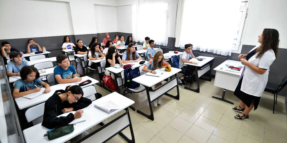 Sınıf ortamında, beyaz önlüklü bir kadın öğretmenin ayakta ders anlattığı, öğrencilerin ise sıralarında oturarak not aldığı bir eğitim merkezi sınıfı.