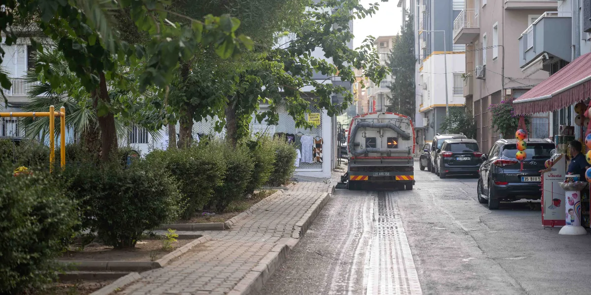 Belediyeye ait bir yol süpürme aracı, yerleşim yerindeki bir sokakta temizlik çalışması yapıyor. Aracın önünde park etmiş otomobillerin bulunduğu yolun kenarında, ağaçlıklı bir park alanı ve sağ tarafta dükkanlar yer alıyor.