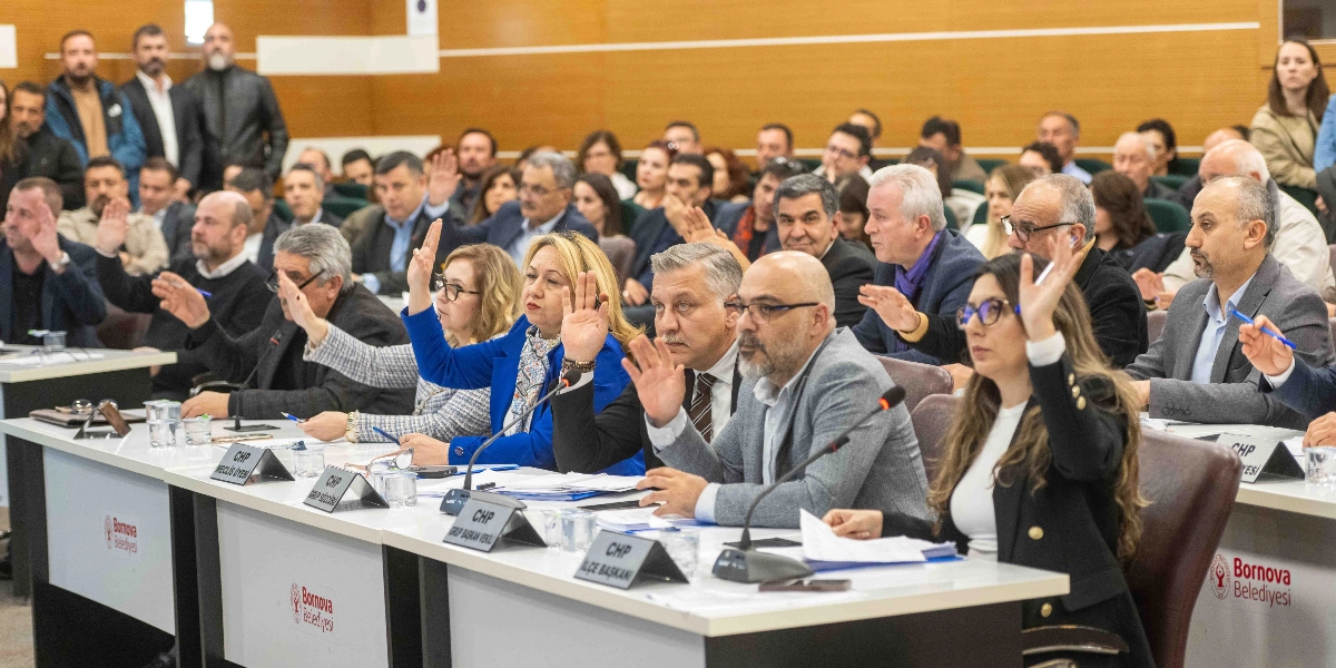 Bornova Belediyesi meclis salonunda, ön masalarda oturan meclis üyelerinin bir oylama sırasında ellerini kaldırarak oy kullandığı toplantı anı. Masaların üzerinde Bornova Belediyesi logoları ve CHP grup üyelerini belirten isimlikler yer alıyor.