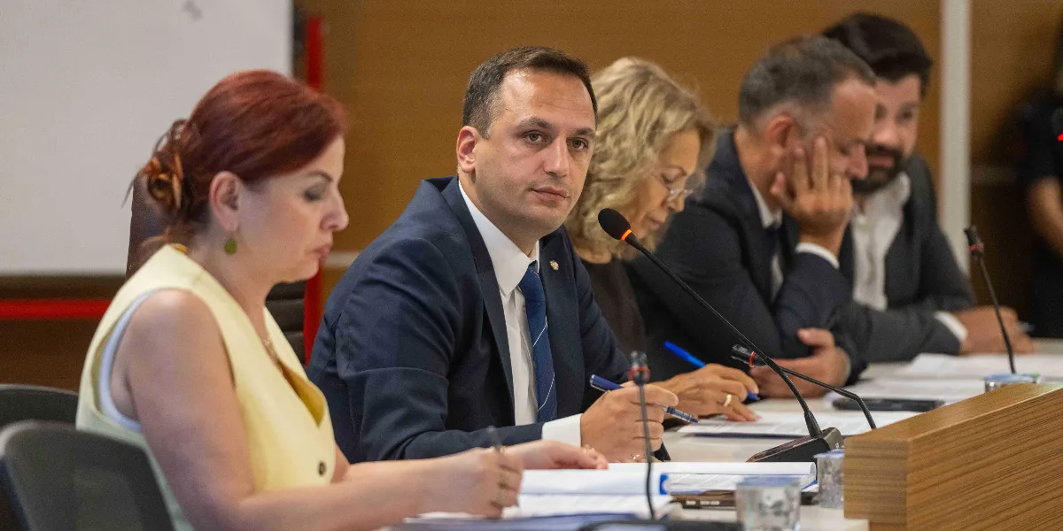 Toplantı masasında oturan, önlerinde evraklar ve mikrofonlar bulunan bir grup yetkili. Ön planda sarı bluzlu bir kadın ve yanında lacivert takım elbiseli, mavi kravatlı bir erkek dikkatle dinliyor veya not alıyor. Arka planda ise diğer katılımcılar toplantıyı takip ediyor.