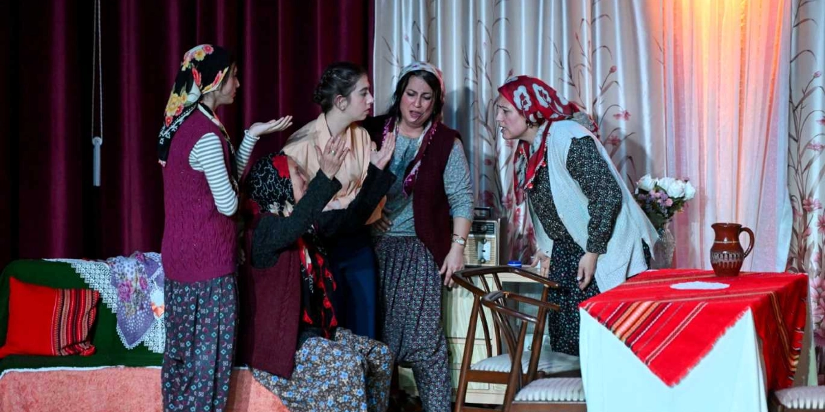 Bir tiyatro sahnesinde, geleneksel kıyafetler giymiş beş kadın duygusal ve gergin bir tartışma anında bir arada duruyor. Solda, başında çiçekli bir yazma olan genç bir kadın ellerini açarak bir şeyler anlatıyor. Onun yanında, sırtı dönük oturan yaşlı bir kadın ellerini havaya kaldırarak konuşuyor. Ortada, üzgün ve ağlamaklı bir ifadeyle duran bir kadın ile yanında ona bakan genç bir kadın bulunuyor. Sağ tarafta ise kırmızı desenli bir başörtüsü takan başka bir kadın, karşısındakine hararetli bir şekilde bir şeyler söylüyor. Arka planda perdeler, önde ise üzerinde kırmızı bir örtü ve toprak bir sürahi bulunan bir masa yer alıyor.