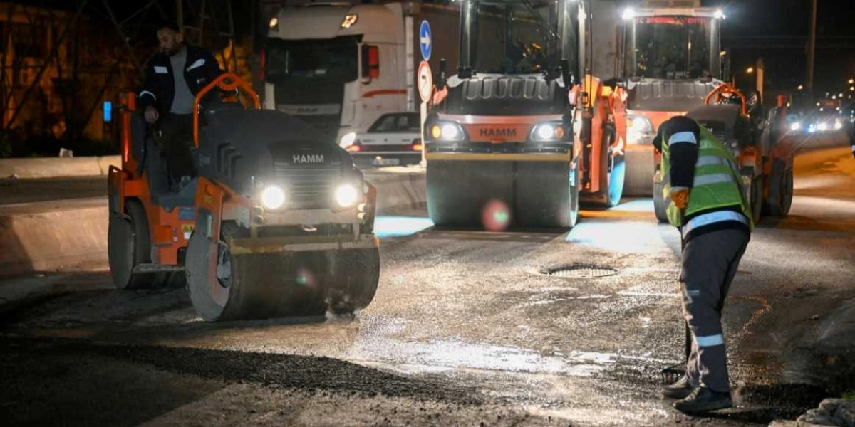 Gece vakti Bornova'da devam eden asfalt serim çalışmasında, üzerinde Hamm marka silindirlerin bulunduğu yolda işçiler görev yapıyor. Reflektörlü yelek giyen bir işçi elindeki tırmıkla asfaltı düzeltirken, diğer işçiler silindirleri kullanıyor. Arka planda ise trafik akışının olduğu yolda park halindeki araçlar ve çalışma ışıkları görülüyor.