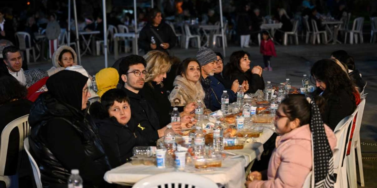 Açık havada, akşam saatlerinde kurulan uzun bir masada iftar yapan insanlar. Masanın üzerinde plastik su şişeleri, yemek kapları ve çeşitli yiyecekler bulunuyor. Farklı yaş gruplarından kadın, erkek ve çocukların bir arada oturduğu masada, katılımcılar yemeklerini yerken veya sohbet ederken görülüyor. Arka planda ise benzer şekilde kurulmuş başka masalar ve insanlar yer alıyor.