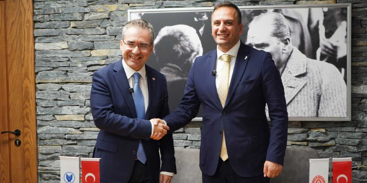 Takım elbiseli iki erkek, taş duvarlı bir odada Mustafa Kemal Atatürk'ün portresinin önünde el sıkışıyor. Soldaki kişi mavi kravatlı, sağdaki kişi ise sarı desenli kravatlıdır. Masanın üzerinde kurum logoları ve Türk bayrakları yer almaktadır.