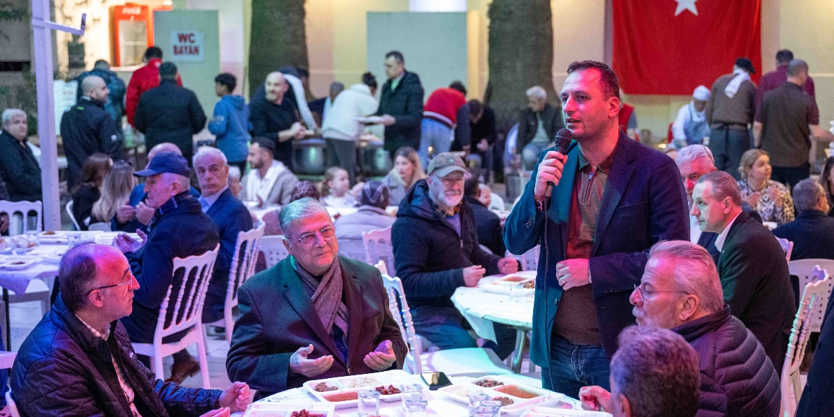 Bornova Belediye Başkanı Ömer Eşki, kalabalık bir iftar sofrasında elinde mikrofonla konuşma yapıyor. Başkanın önündeki masada oturan katılımcılar ellerini dua eder gibi açmış dinlerken, arka planda Türk bayrağının asılı olduğu açık hava alanında çok sayıda kişi yemek yiyor.