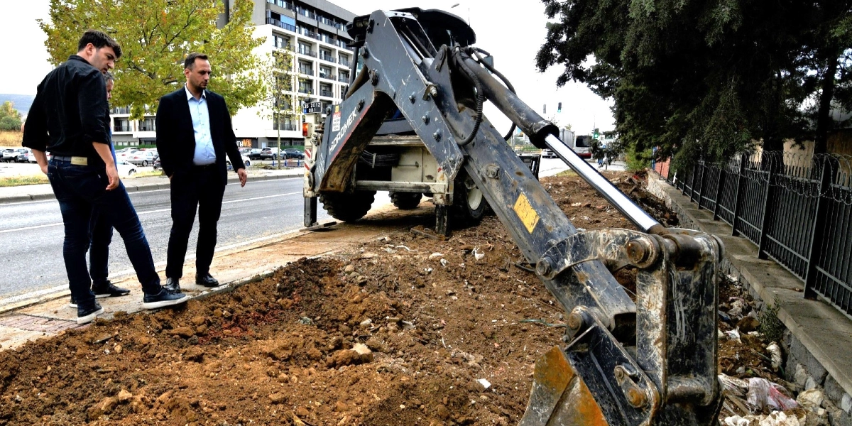 Siyah ceketli iki erkek, yol kenarındaki toprak alanda çalışan Hidromek marka bir iş makinesini inceliyor. Arka planda modern bir bina ve trafik akışının olduğu bir yol bulunuyor.