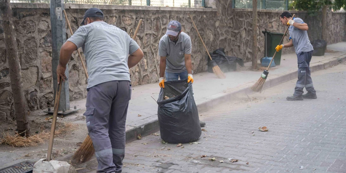 Bornova Belediyesi üniformalı üç temizlik işçisi, taş duvarlı bir yol kenarında süpürge ve çöp poşetleriyle temizlik yapıyor. İşçilerden biri ortada durarak elindeki çöp poşetini açık tutarken, diğer ikisi ellerindeki süpürgelerle kaldırım kenarındaki toz ve yaprakları temizliyor.