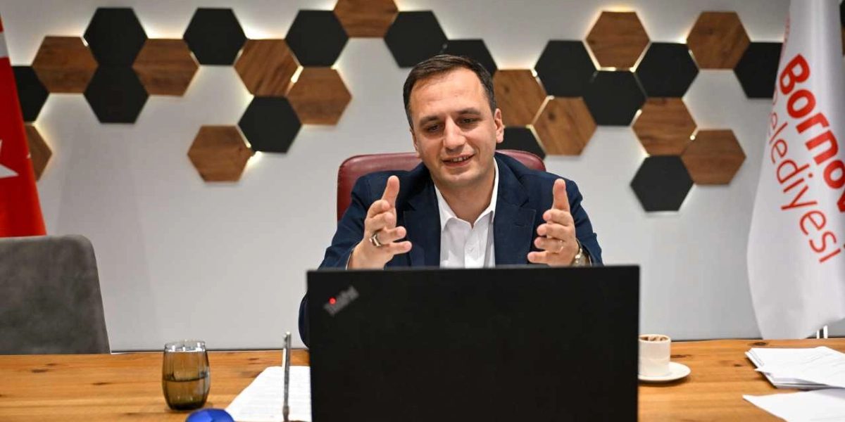 Koyu renk ceket ve beyaz gömlek giymiş bir erkek, ahşap bir masa başında oturarak dizüstü bilgisayardan çevrim içi bir toplantıya katılıyor. Elleriyle konuşmasına eşlik eden jestler yapan kişinin arkasında, Bornova Belediyesi logosunun bulunduğu bir bayrak ve altıgen desenli, ışıklandırılmış dekoratif bir duvar paneli yer alıyor. Masanın üzerinde bir bardak su, bir fincan kahve ve bazı evraklar bulunuyor.