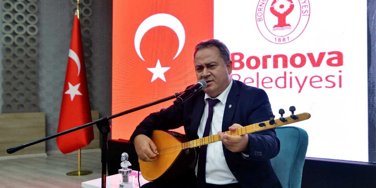 Koyu renk takım elbiseli ve kravatlı bir adam, mikrofon önünde oturarak bağlama çalıyor. Arka planda Türk bayrağı ve üzerinde Bornova Belediyesi logosu ile yazısı bulunan büyük bir dijital ekran yer alıyor.