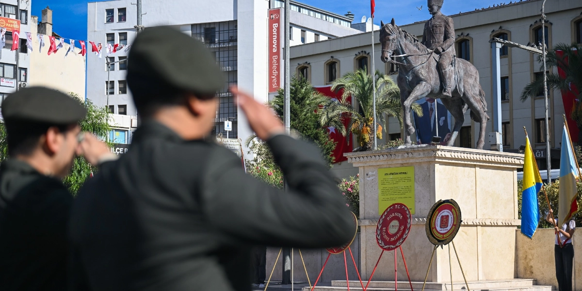 Bornova'da düzenlenen 19 Eylül Gaziler Günü töreninde, ön planda selam duran üniformalı askerler, arka planda ise üzerinde Atatürk'ün atlı heykeli bulunan kaide yer alıyor. Kaidenin önünde Bornova Belediyesi ve Türkiye Muharip Gaziler Derneği'ne ait çelenkler bulunurken, çevrede Türk bayrakları ve belediye binası görülüyor.