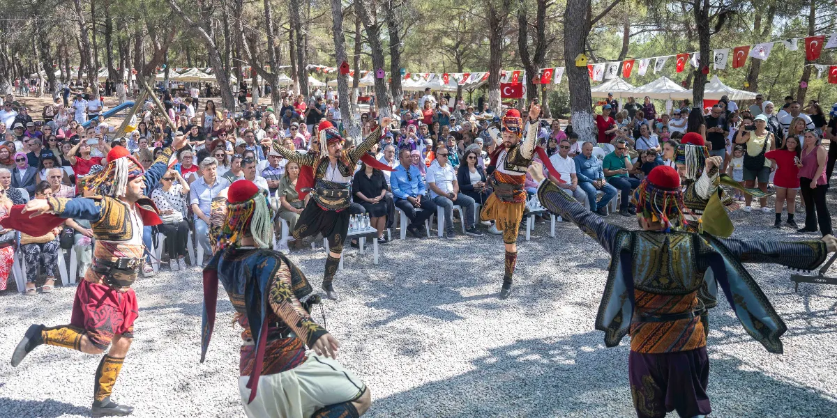 Yakaköy’deki bir ağaçlık alanda, geleneksel kıyafetler giymiş dört kişilik bir halk oyunları ekibi gösteri yaparken, onları izleyen kalabalık bir grup izleyici sandalyelerde oturuyor. Dansçılar, işlemeli yelekler, şalvarlar ve başlıklar içinde kollarını açarak hareket ederken, arka planda ağaçlara asılmış Türk bayrakları ve şenlik alanındaki beyaz çadırlar görülüyor.