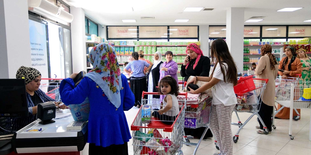 Bornova Belediyesi'ne ait bir marketin kasa bölümünde, alışveriş yapan kadınlar ve çocuklar görülüyor. Ön planda mavi kıyafetli ve çiçekli başörtülü bir kadın kasada işlem yaparken, yanında alışveriş arabasında küçük bir çocuk oturuyor. Arka planda ise raflarla dolu marketin içinde alışveriş yapan, farklı kıyafet ve başörtüleri olan kadınlar bulunuyor. Bazı kadınlar alışveriş arabalarını doldurmuş beklerken, bazıları telefonla konuşuyor veya ürün seçiyor. Marketin camlarında Bornova Belediyesi logoları ve sosyal destek mesajları yer alıyor.