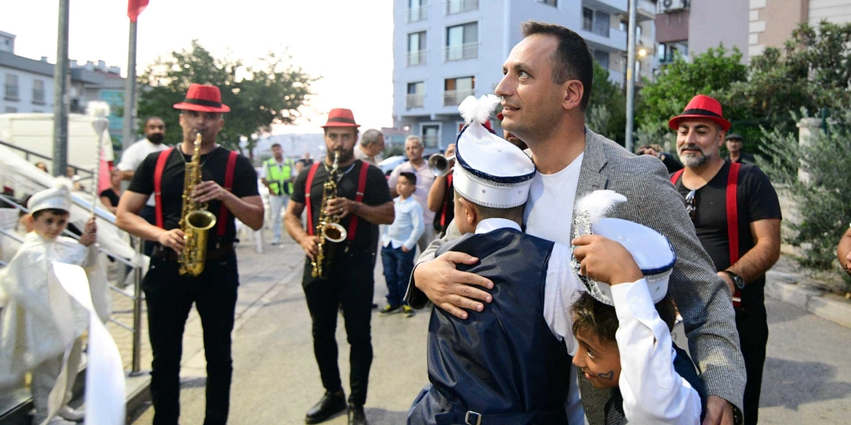 Sünnet kıyafetleri giymiş iki çocuğu kucaklayan bir adam ve arkalarında saksafon çalan, kırmızı şapkalı ve kırmızı askılı müzisyenlerin yer aldığı bir sokak kutlaması.