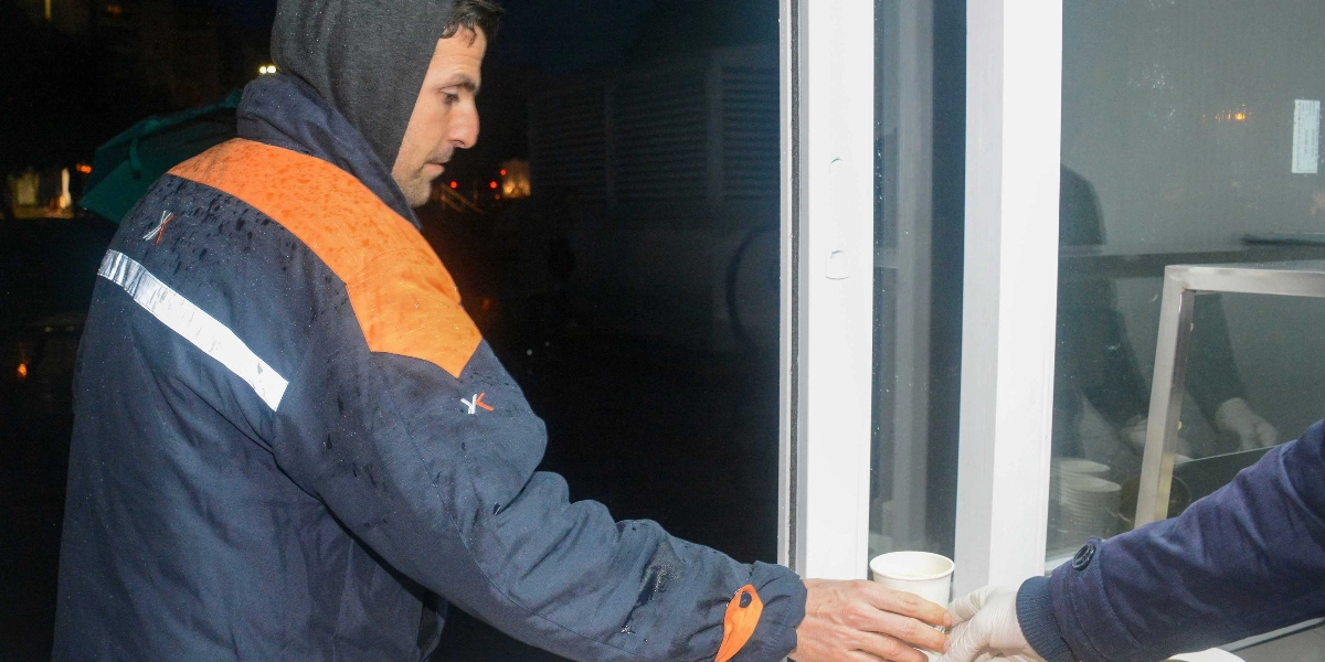 Kapüşonlu koyu renkli bir mont giyen bir çalışan, beyaz eldivenli bir kişiden karton bardakta sıcak bir içecek alıyor.