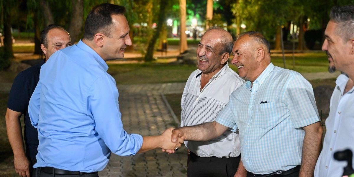 Mavi gömlekli bir erkek, akşam saatlerinde bir parkta orta yaşlı bir adamla el sıkışırken gülümsüyor. Çevredeki diğer iki erkek de bu anı gülümseyerek izliyor.