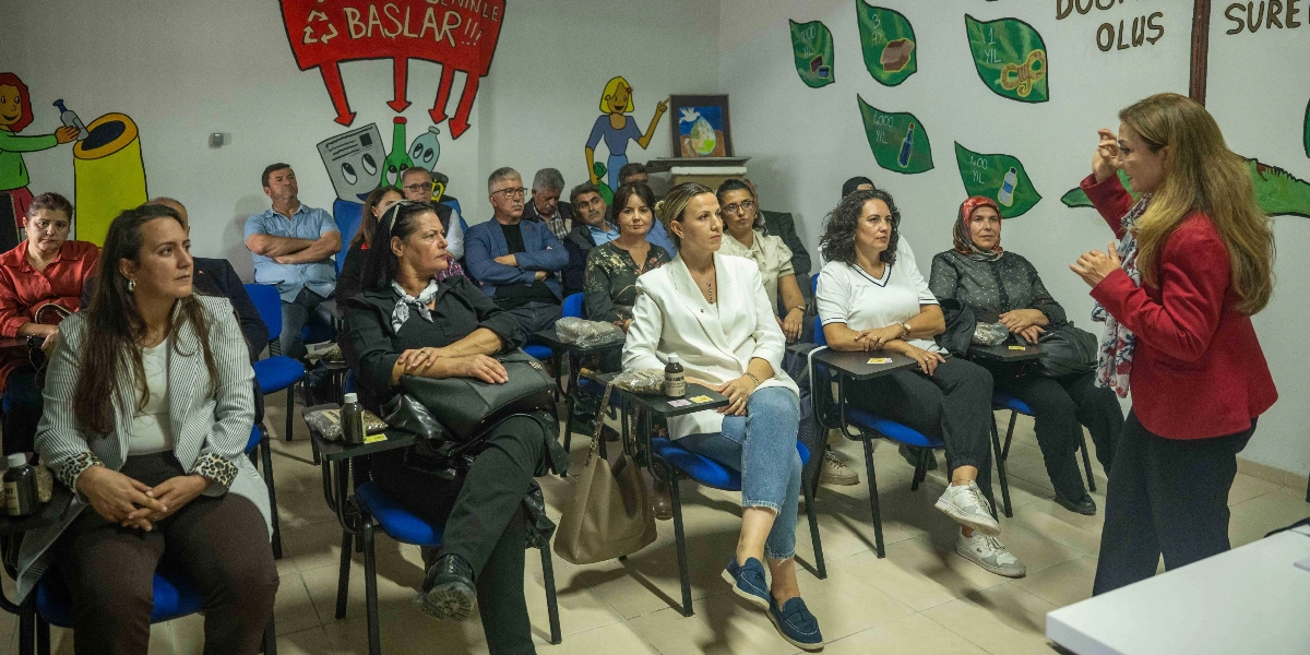 Sınıf ortamında, duvarlarında sıfır atık temalı çizimlerin ve bilgilendirici yazıların bulunduğu bir odada, kırmızı ceketli bir kadın ayakta sunum yaparken, katılımcı grubu onu dikkatle dinliyor. Katılımcılar mavi sandalyelerde oturuyor ve önlerindeki sıralarda küçük numune paketleri bulunuyor.