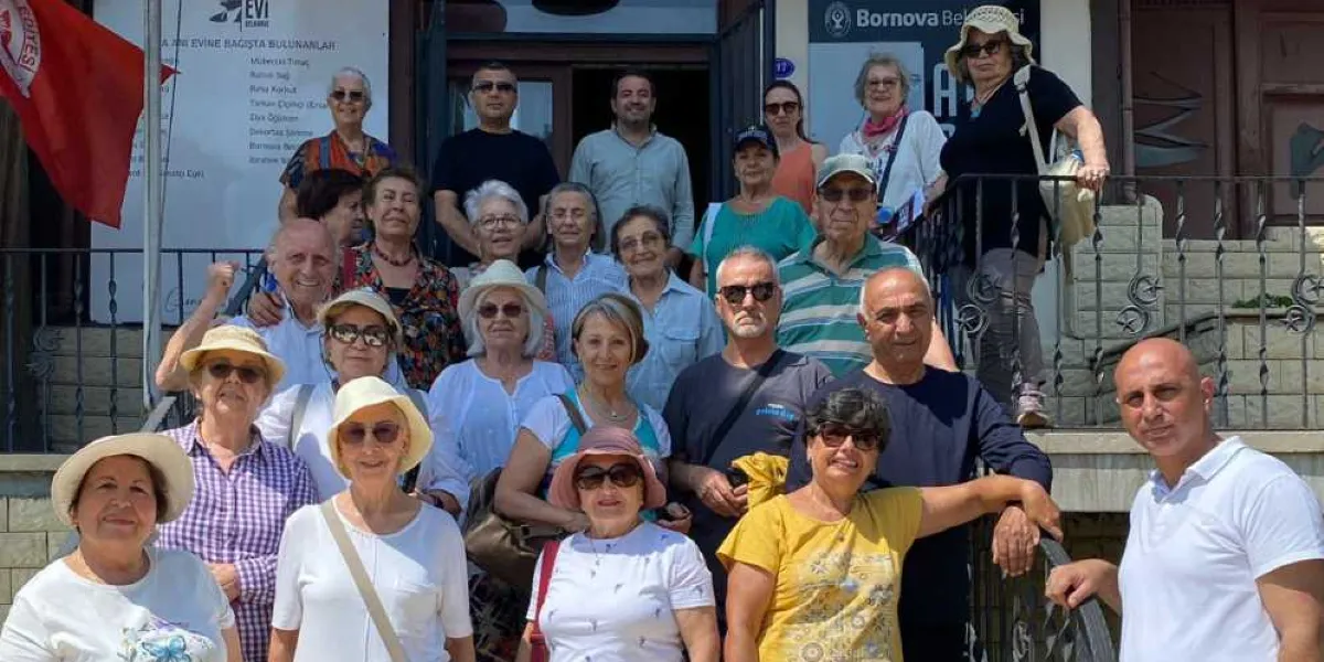 Bornova Belediyesi binasının girişindeki merdivenlerde poz veren, farklı yaş gruplarından oluşan kalabalık bir grup insan. Çoğunluğu güneş gözlüğü ve şapka takan katılımcılar, güneşli bir günde dış mekanda yan yana durarak gülümsüyor. Arka planda Bornova Belediyesi yazılı bir tabela ve üzerinde isimlerin yer aldığı bir bilgilendirme panosu ile kırmızı bir bayrak görülüyor.
