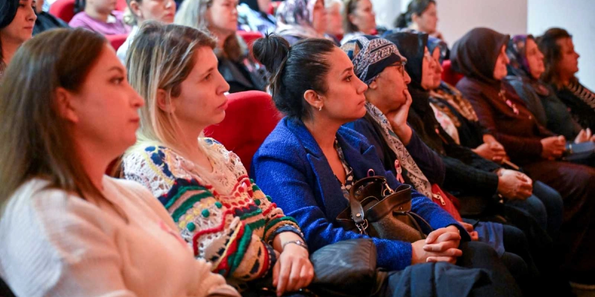 Konferans salonunda yan yana oturmuş, dikkatle dinleyen kadınlardan oluşan bir grup. Ön planda beyaz kazaklı, renkli desenli hırkalı ve mavi ceketli kadınlar yer alıyor. Bazı katılımcıların yakalarında meme kanseri farkındalığını simgeleyen pembe kurdeleler bulunuyor. Katılımcıların bir kısmı başörtülü, bir kısmı ise açık. Hepsi sahneye doğru dönmüş, odaklanmış bir şekilde oturuyor.